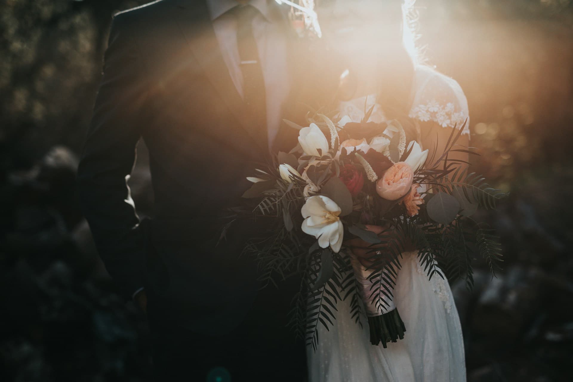 Bride and groom stand side by side.  Bride is carrying a large bouquet of white, pink, and red flowers.  The image background is dark and softly muted, but the sun is shining from behind and lighting the bride's hair.